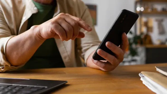Persona usando un smartphone en un escritorio junto a un portátil para representar la conectividad y banca digital.