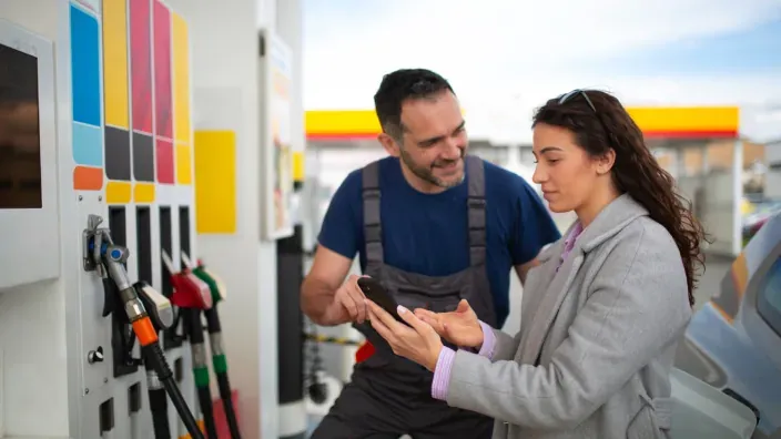 Empleado de una gasolinera ayudando a una mujer que consulta el móvil junto a un surtidor