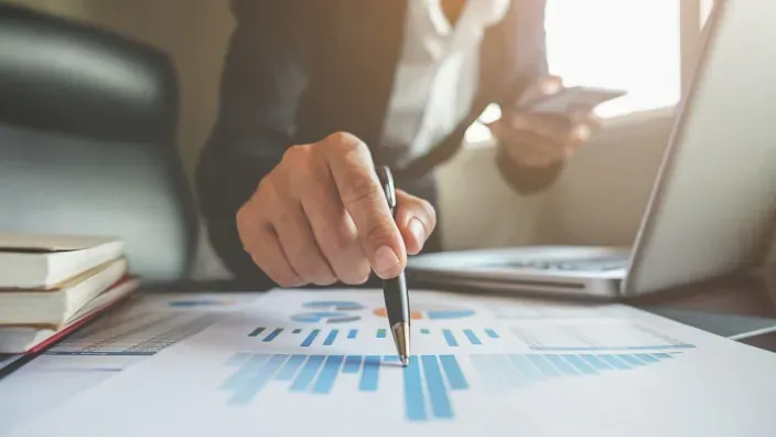 Persona señalando un gráfico financiero impreso sobre una mesa con un portátil y un móvil al fondo
