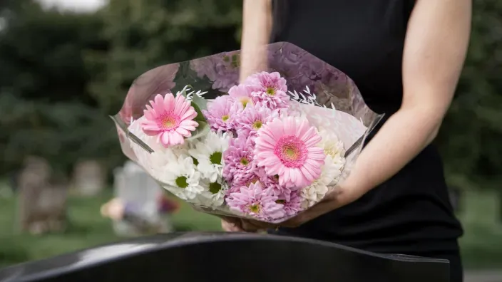 Manos de una persona sosteniendo un ramo de flores frente a una tumba, en señal de recuerdo y homenaje.