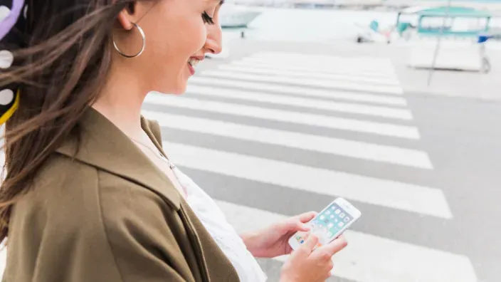 Mujer joven sonriente usando un teléfono móvil smartphone en un paso de cebra en la calle.