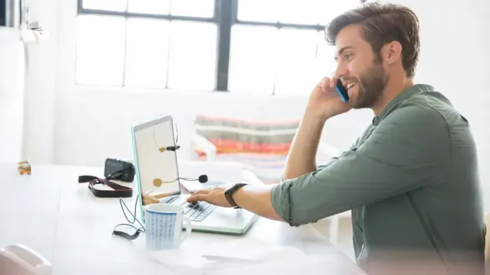 Hombre sonriente teletrabajando en una oficina. Habla por el móvil mientras usa su ordenador portátil para gestionar su negocio.