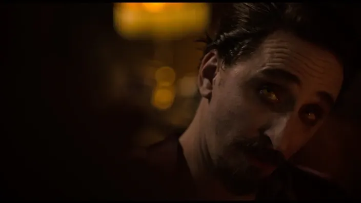 Primer plano de un hombre con una mirada siniestra y ojos amarillos brillantes en una habitación oscura, creando una atmósfera de terror.