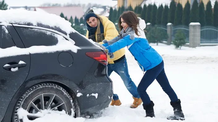 Pareja empujando un coche atrapado en la nieve durante una nevada.