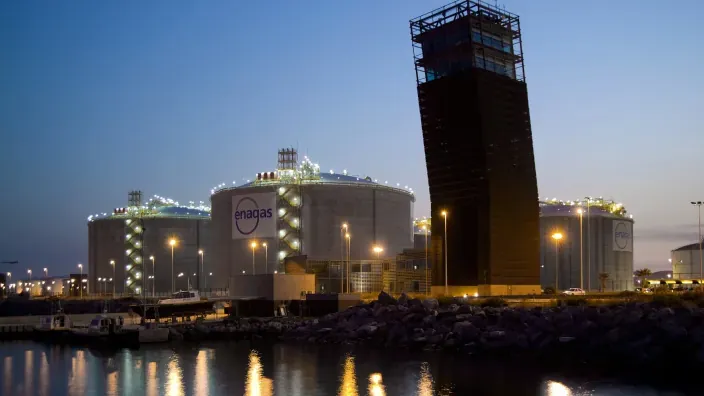 Planta regasificadora de Enagás iluminada de noche en zona portuaria con depósitos de gas y torre industrial junto al mar.