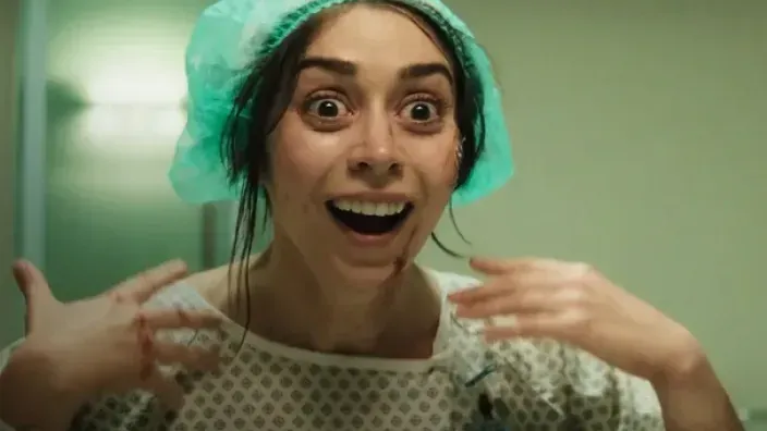 Mujer con gorro quirúrgico sonriente y manchada de sangre en una escena tensa dentro de un hospital.