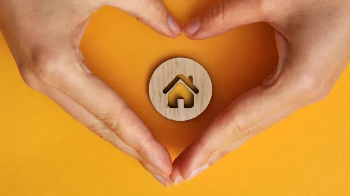 Manos formando un corazón alrededor de un icono de casa para representar la protección y el seguro de hogar.