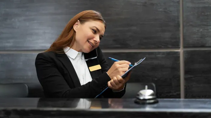 Recepcionista hablando por teléfono mientras escribe en un portapapeles detrás de un mostrador.