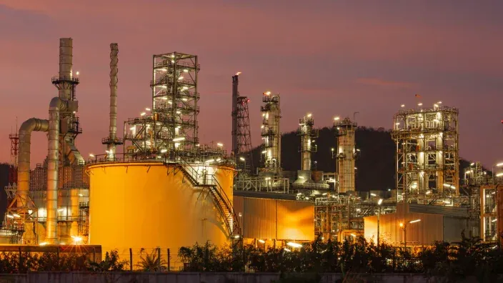Complejo industrial de una refinería de petróleo con torres y tanques iluminados al atardecer.