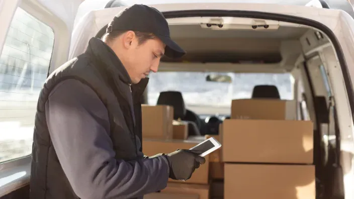Repartidor revisando el móvil junto a una furgoneta de reparto llena de paquetes.