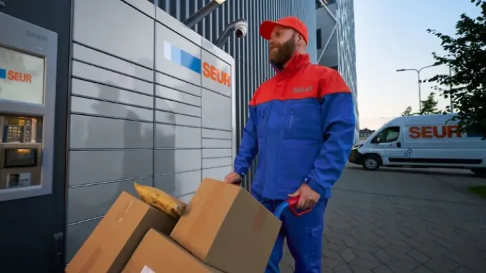 Repartidor de SEUR transportando varios paquetes frente a un punto de recogida automático de la empresa.