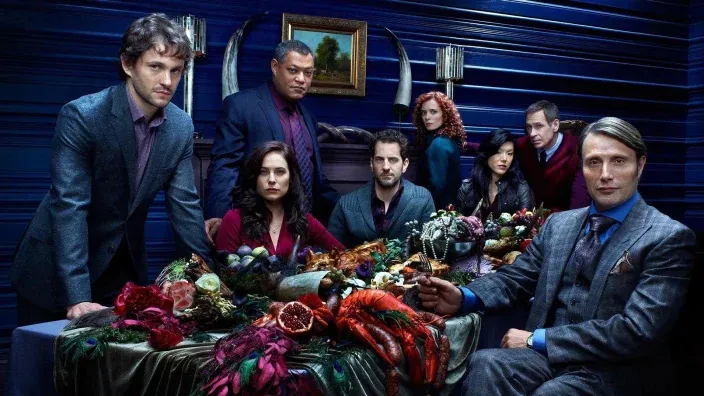 Imagen promocional con todo el elenco de actores y actrices de la serie Hannibal sentados alrededor de una mesa con un banquete de carne.