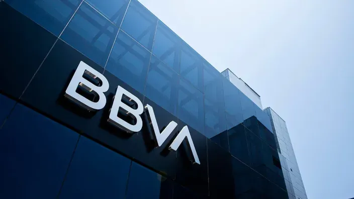 Logotipo de BBVA en letras blancas sobre la fachada de cristal oscuro de un edificio de la entidad bancaria, visto desde abajo.