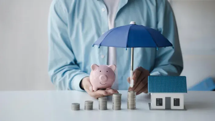 Concepto de protección financiera con paraguas cubriendo hucha cerdito, monedas y maqueta de casa.