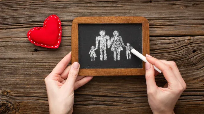 Manos dibujando siluetas de familia en pizarra negra junto a un corazón rojo de fieltro.