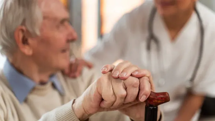 Cuidadora brindando apoyo a un anciano que sujeta un bastón, simbolizando asistencia médica y cuidados a la tercera edad.