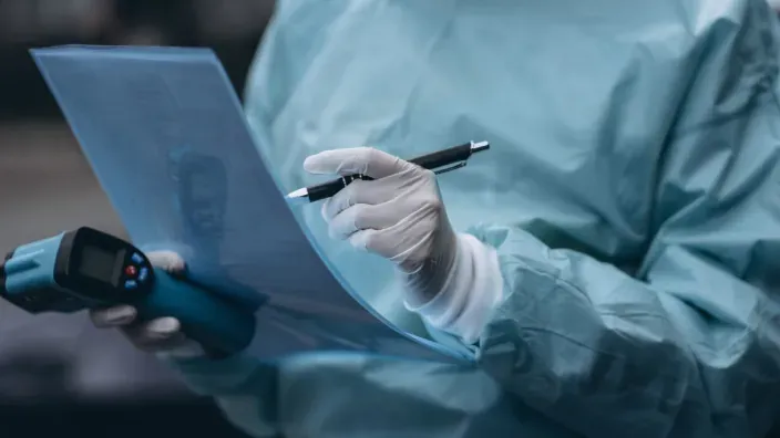 Profesional sanitario revisando documentos médicos con guantes y dispositivo electrónico.