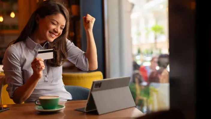 Mujer celebrando con su tarjeta bancaria frente a una tablet en una cafetería.