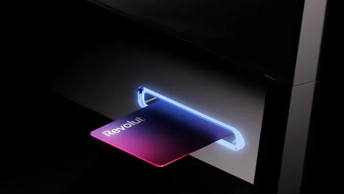 Tarjeta de débito del neobanco Revolut saliendo de la ranura iluminada de un cajero automático.
