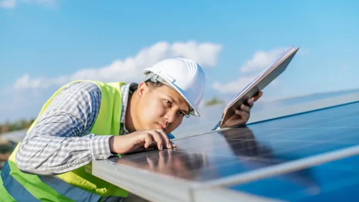 Técnico con casco y chaleco reflectante revisando un panel solar mientras sostiene una tablet