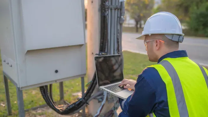 Técnico de fibra óptica usando un portátil para mantenimiento de red de telecomunicaciones en el exterior.