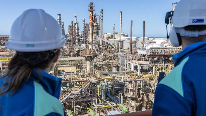 Trabajadores con equipos de seguridad caminando frente a una refinería industrial de energía y combustibles.