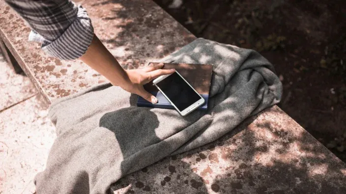 Persona colocando la mano sobre un teléfono móvil situado encima de varias prendas en un banco de piedra