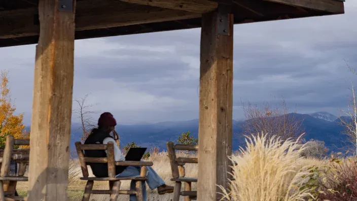 Mujer teletrabajando con su portátil desde un porche de madera en la montaña para representar la conectividad rural.