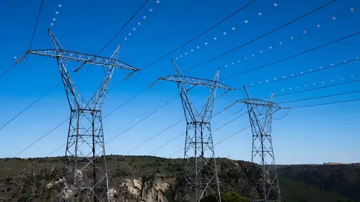 Torres de alta tensión transportando electricidad sobre un paisaje montañoso bajo un cielo azul despejado.