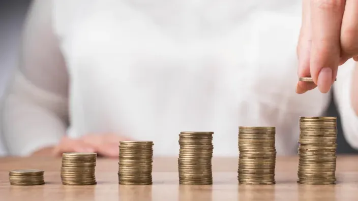 Mano de una persona colocando una moneda sobre una pila creciente de monedas.