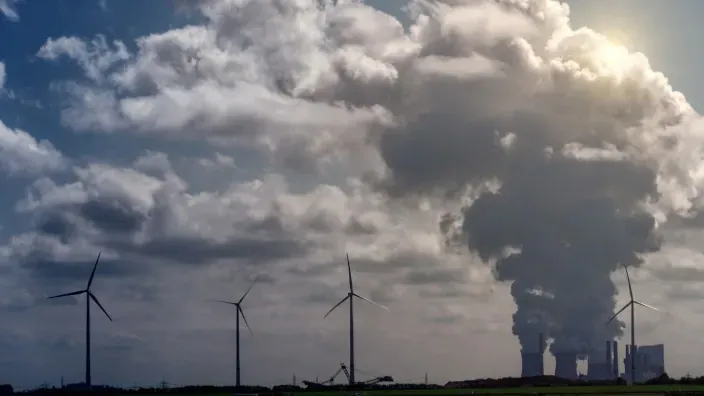 Imagen de torres de refrigeración expulsando vapor, con aerogeneradores al fondo bajo un cielo nublado.