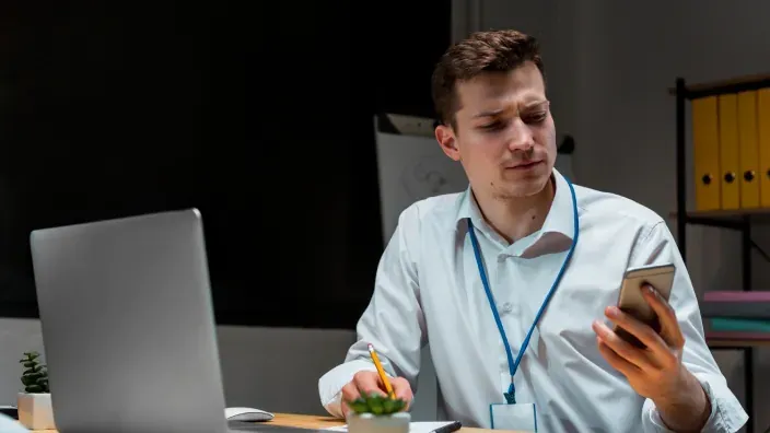 Trabajador autónomo consultando su teléfono móvil con expresión de duda frente al ordenador.