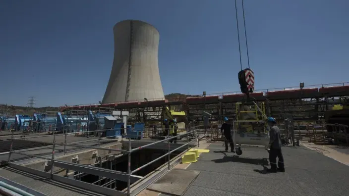 Operarios trabajando en las instalaciones exteriores de una central nuclear con una torre de refrigeración al fondo.