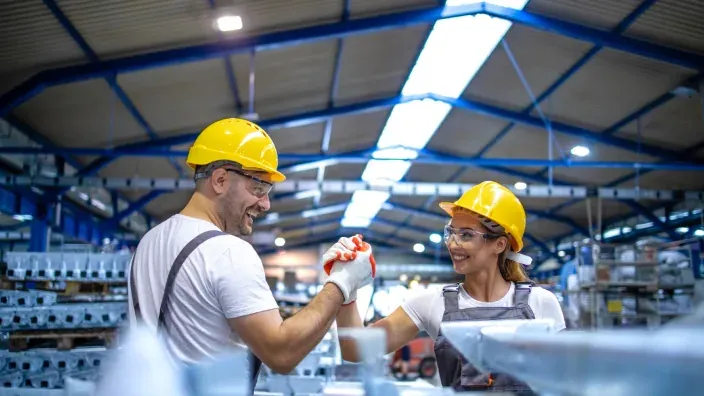 Dos operarios con casco amarillo chocan las manos en una nave industrial.
