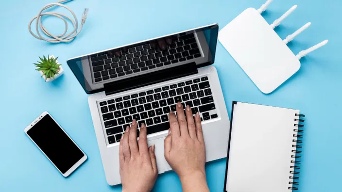 Persona escribiendo en un ordenador portátil junto a un router WiFi, un móvil y un cuaderno sobre fondo azul, representando conexión a internet.