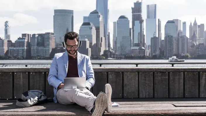 Hombre con auriculares y portátil frente al skyline de una ciudad para representar el teletrabajo y la conectividad global.