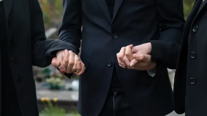 Tres personas vestidas de negro se toman de las manos en señal de apoyo durante un funeral.