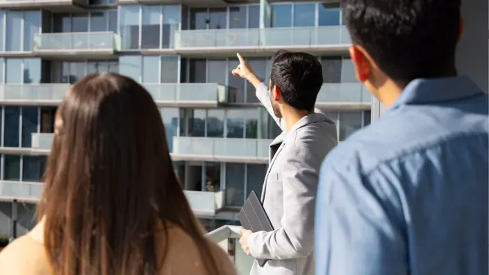 Una pareja joven visita un piso para comprar mientras el agente inmobiliario enseña las vistas desde la terraza.