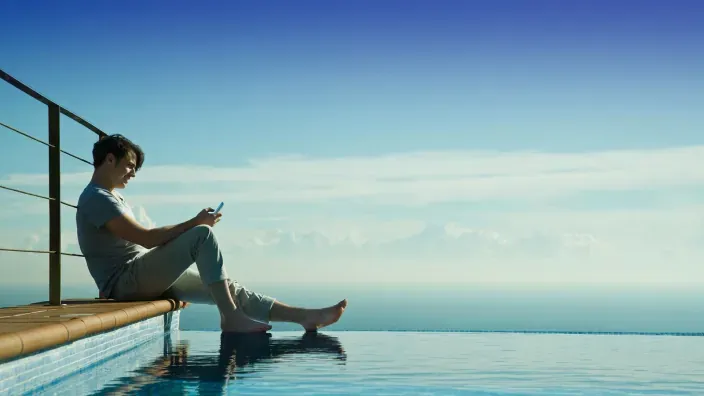 Hombre usando su smartphone junto a una piscina frente al mar para representar el uso de internet móvil en vacaciones.