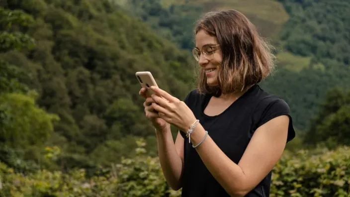 Mujer usando su smartphone en la montaña para representar la conectividad móvil por satélite en entornos rurales.