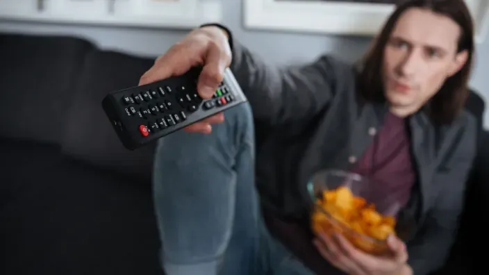 Persona usando mando a distancia y comiendo snacks en el sofá para disfrutar de cine y series en streaming en casa.