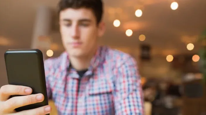 Hombre joven desenfocado al fondo consultando la pantalla de un smartphone negro en primer plano.