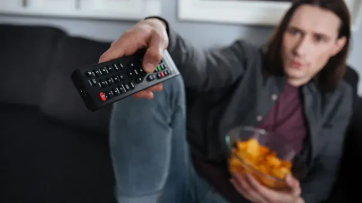 Hombre en el sofá usando el mando a distancia para cambiar de canal mientras come snacks frente al televisor.