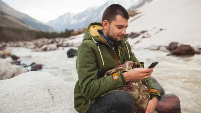 Hombre usando su móvil junto a un río en la montaña para representar la conectividad satelital en zonas remotas.