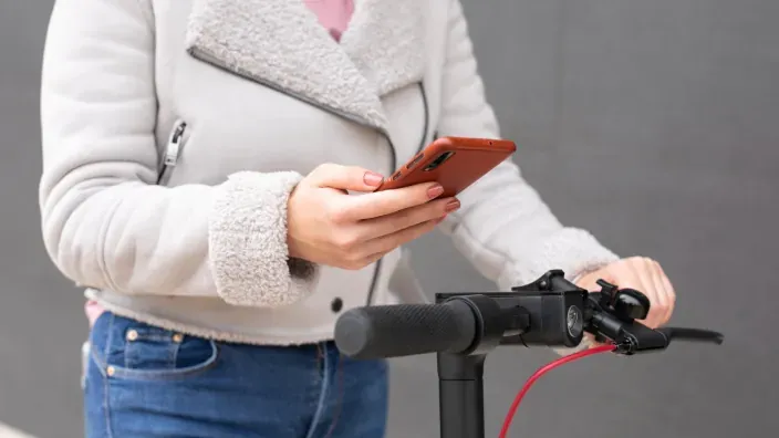 Mujer usando su smartphone junto a un patinete eléctrico, reflejo de la tecnología y movilidad urbana.