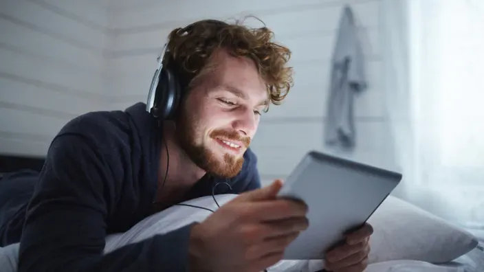 Hombre con auriculares usando una tablet en la cama de un hotel para ver cine y series en streaming.
