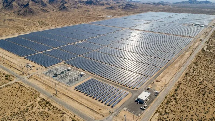 Vista aérea de una gran planta de energía solar fotovoltaica con miles de paneles alineados en una zona desértica bajo la luz del sol.