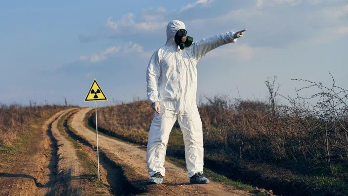 Técnico con traje de protección radiactiva y máscara de gas señalando junto a cartel de peligro nuclear.