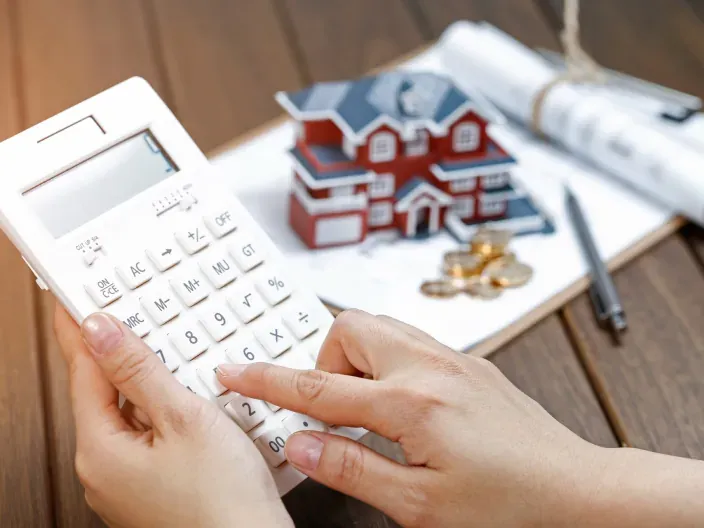 Mano usando calculadora junto a una maqueta de casa, planos y monedas.