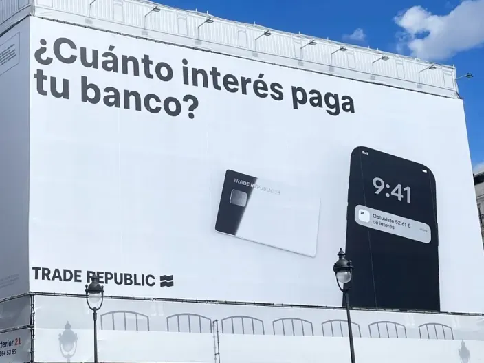 Cartel publicitario de Trade Republic con el mensaje “¿Cuánto interés paga tu banco?” y una tarjeta junto a un móvil mostrando beneficios.